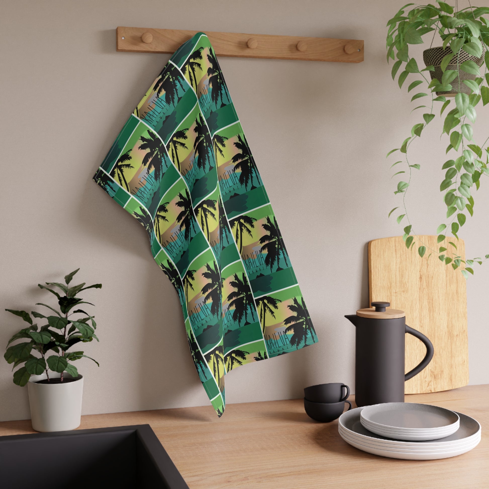 A kitchen towel with a green and black palm tree pattern 'Kailua Hawaii' hanging on a wooden peg rack on a beige wall above a wooden countertop with kitchen items.