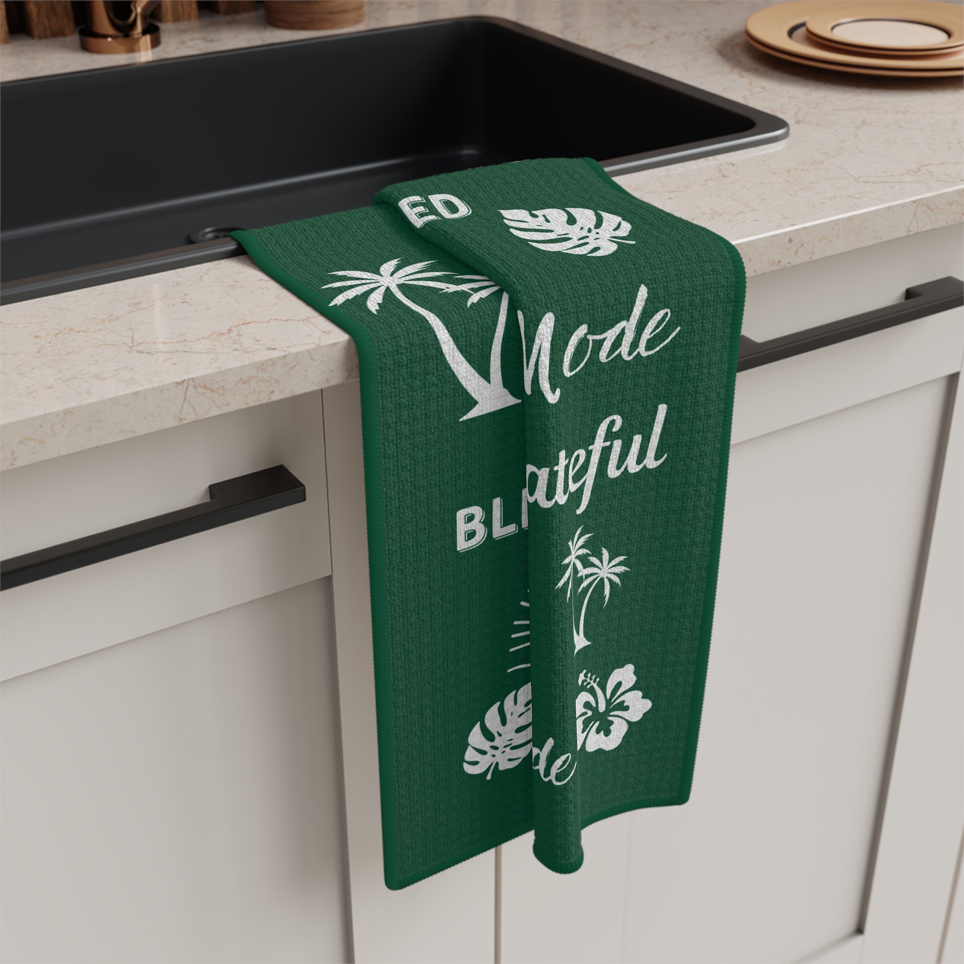 A dark green kitchen towel with white tropical-themed illustrations and repeated phrases including 'Grateful Mahalo Mode,' 'THANKFUL,' 'BLESSED,' and 'Mahalo Mode.' hangs off of kitchen counter.