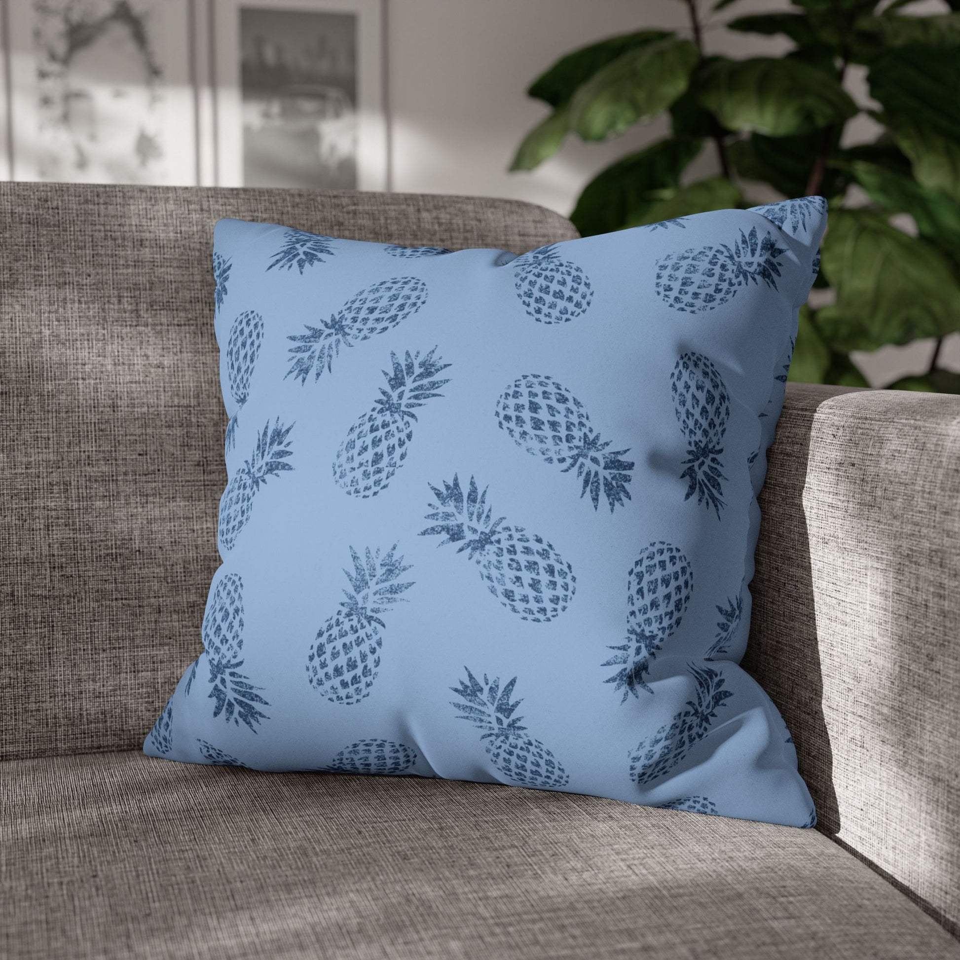A square throw pillow with a blue and navy pineapple pattern, resting on a textured gray fabric sofa with a blurred indoor background.