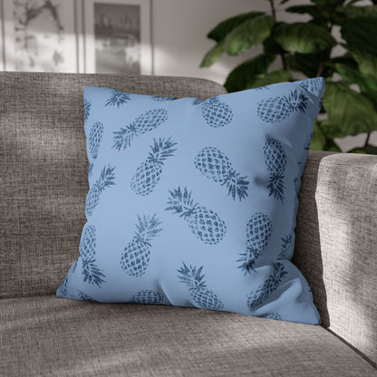 A square throw pillow with a blue and navy pineapple pattern, resting on a textured gray fabric sofa with a blurred indoor background.