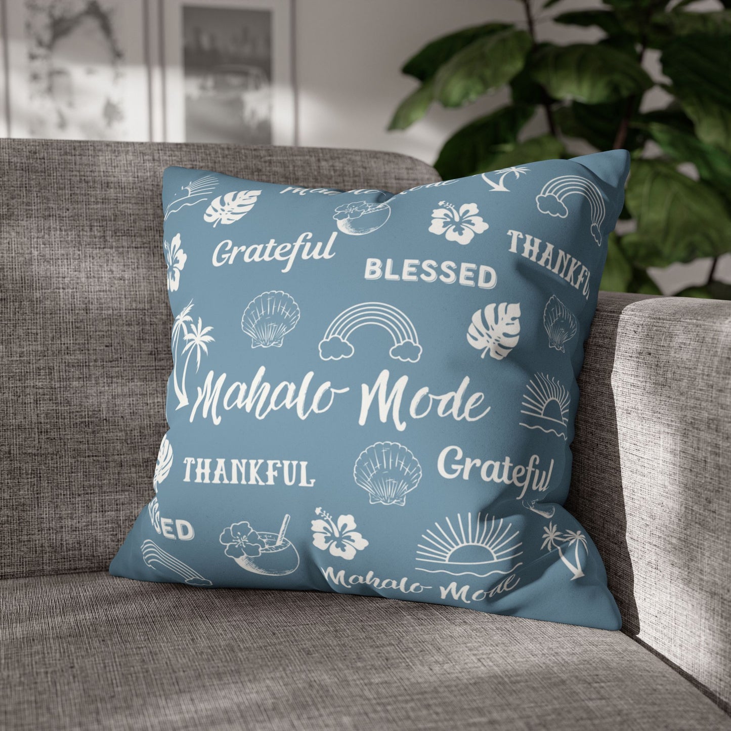 A square throw pillow in dusty blue with white tropical-themed illustrations and words like 'Grateful', 'Blessed', 'Thankful', and 'Mahalo Mode', resting on a gray fabric sofa with a blurred indoor background and plant.