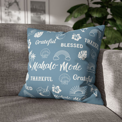 A square throw pillow in dusty blue with white tropical-themed illustrations and words like 'Grateful', 'Blessed', 'Thankful', and 'Mahalo Mode', resting on a gray fabric sofa with a blurred indoor background and plant.