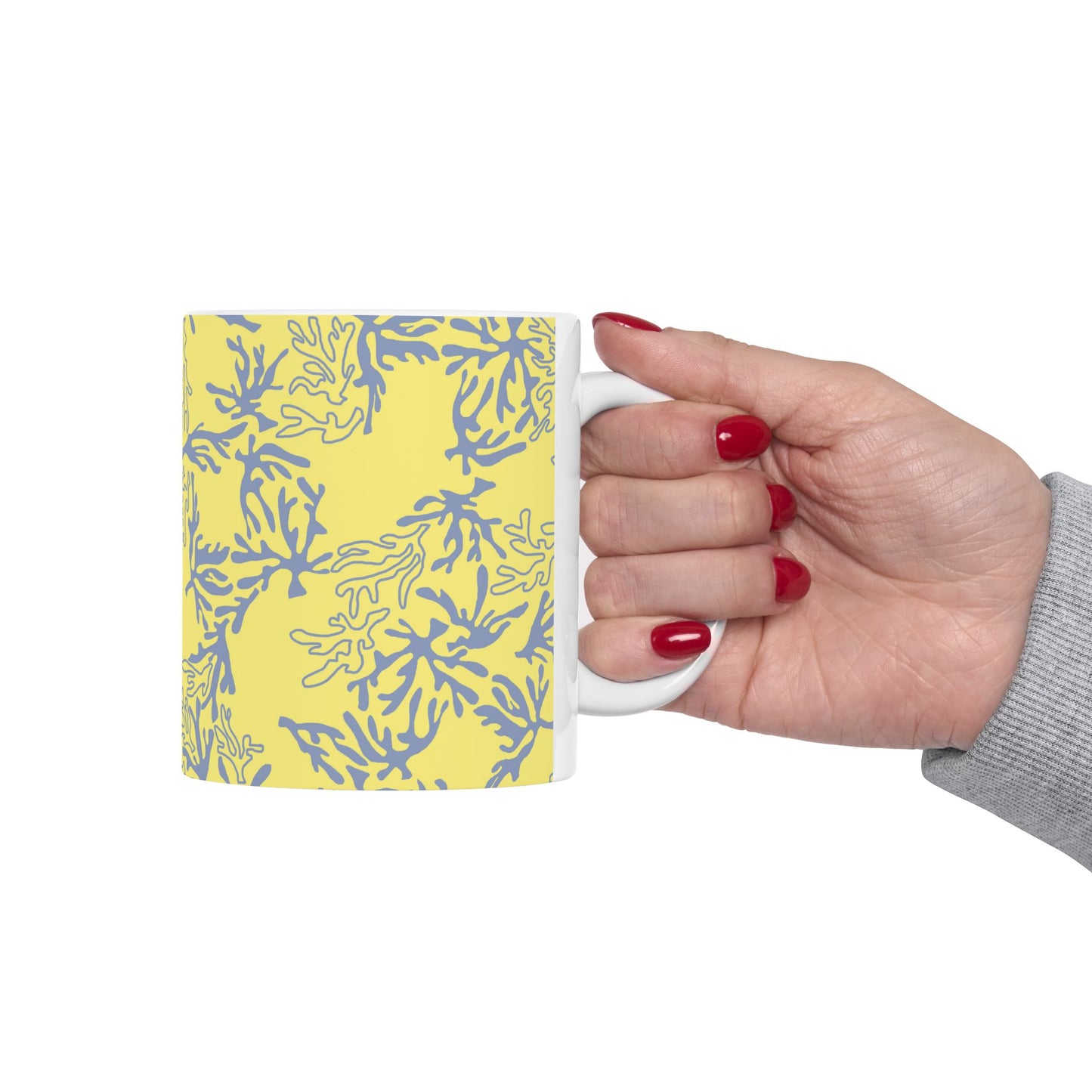 A hand with red nail polish holds a white ceramic mug featuring a yellow and blue coral-like pattern, set against a plain white background.