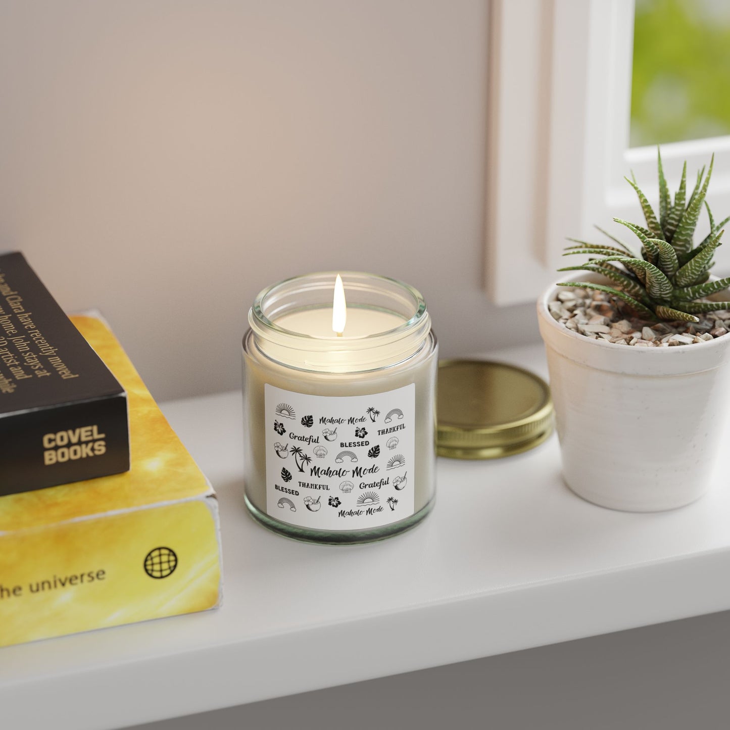 A glass jar candle with a gold metal lid and a white paper label featuring black tropical-themed illustrations and text like 'Mahalo Mode,' 'Grateful,' and 'Blessed,' shown on a table with a plant and books.
