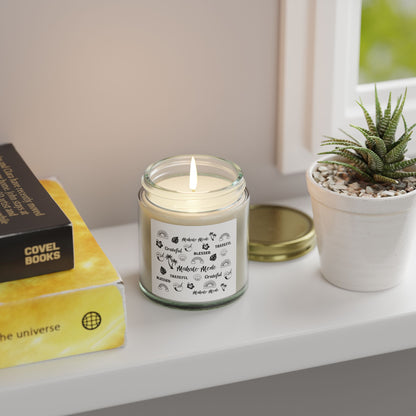 A glass jar candle with a gold metal lid and a white paper label featuring black tropical-themed illustrations and text like 'Mahalo Mode,' 'Grateful,' and 'Blessed,' shown on a table with a plant and books.
