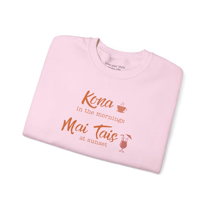 A light pink folded crewneck sweatshirt with a coral orange graphic print that reads 'Kona in the mornings Mai Tais at sunset' with a coffee cup and cocktail glass illustration, shown on a white background.