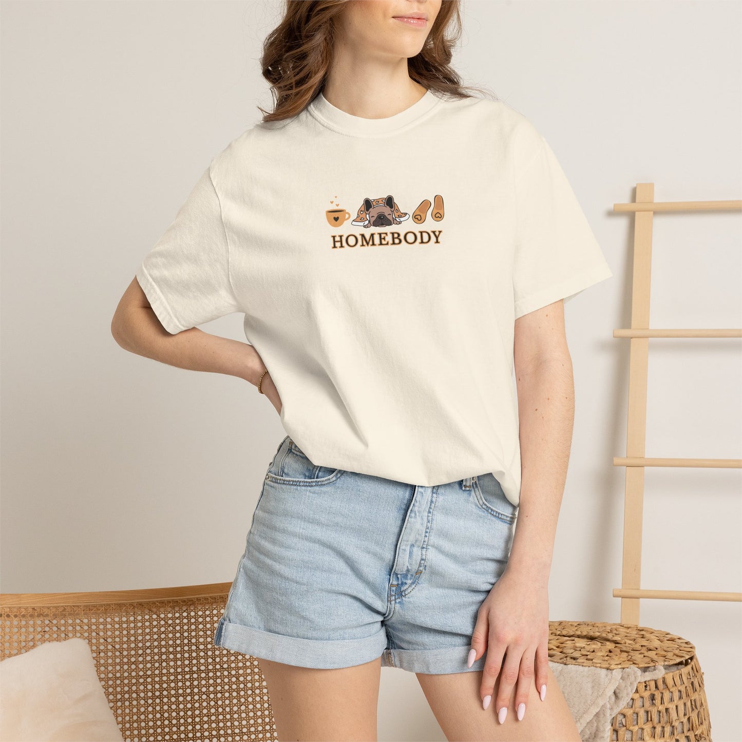 A woman wearing a cream-colored short-sleeve t-shirt with a 'HOMEBODY' graphic on the chest, paired with light blue denim shorts, standing in a minimalist indoor setting.