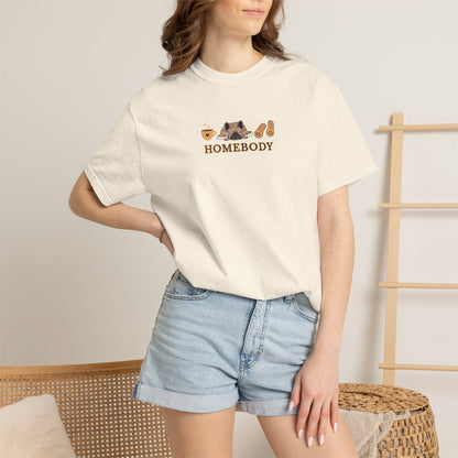 A woman wearing a cream-colored short-sleeve t-shirt with a 'HOMEBODY' graphic on the chest, paired with light blue denim shorts, standing in a minimalist indoor setting.