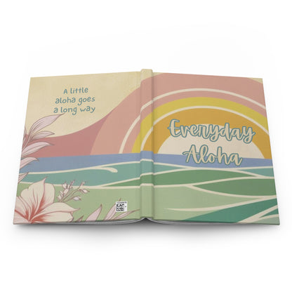 An open hardcover book with illustrated cover pages showing a pastel sunset, ocean waves, tropical flowers, and the text 'A little aloha goes a long way' and 'Everyday Aloha'.