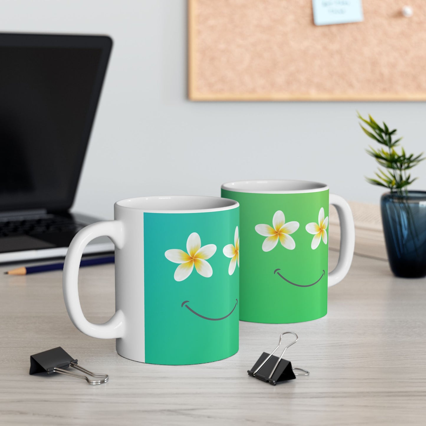 Two ceramic mugs on a light wood desk in a home office setting. The left mug is white with a vertical teal band and a smiling face made of white plumeria flowers for eyes and a curved smile.
