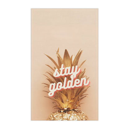 A tea towel featuring a golden pineapple on a warm beige background with the phrase 'stay golden' in white script outlined in pink, shown against a plain white backdrop.