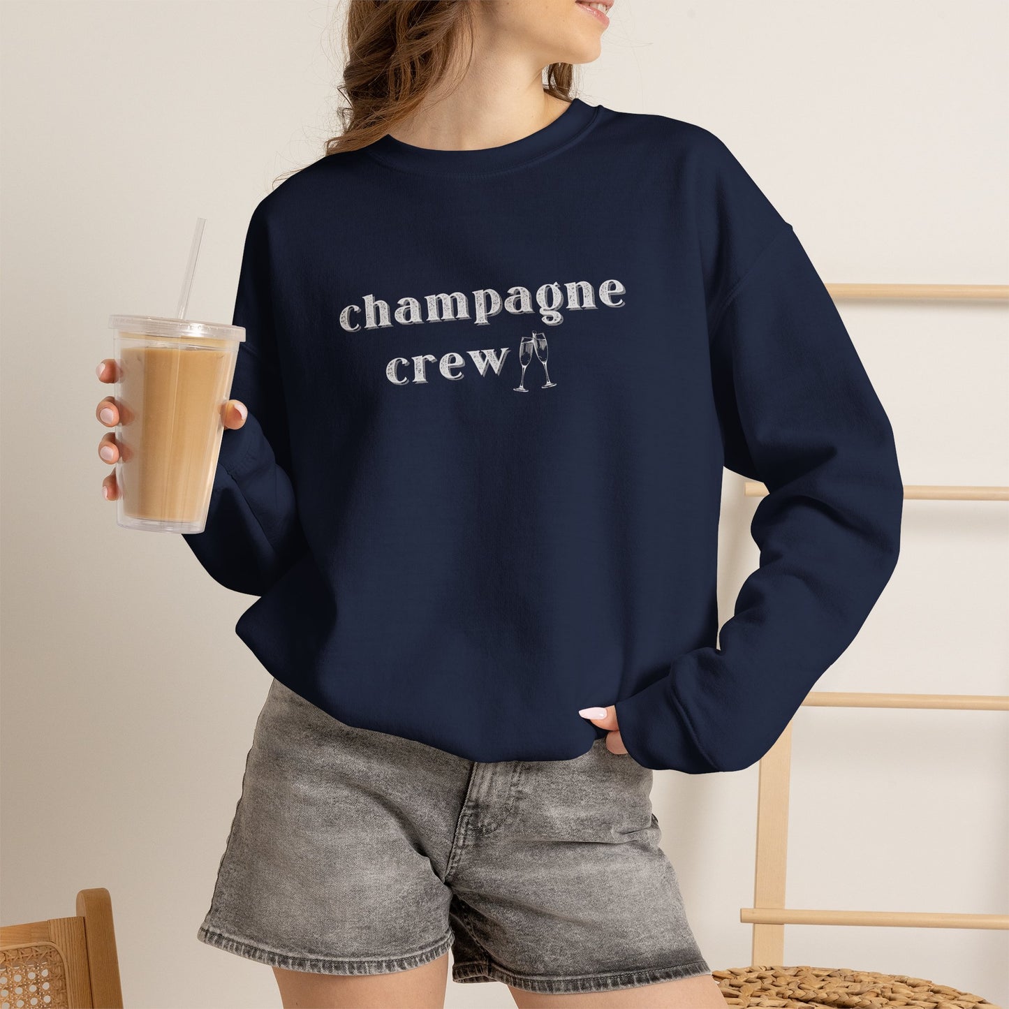Woman wearing a navy graphic sweatshirt with text 'champagne crew' in white letters holding an iced coffee
