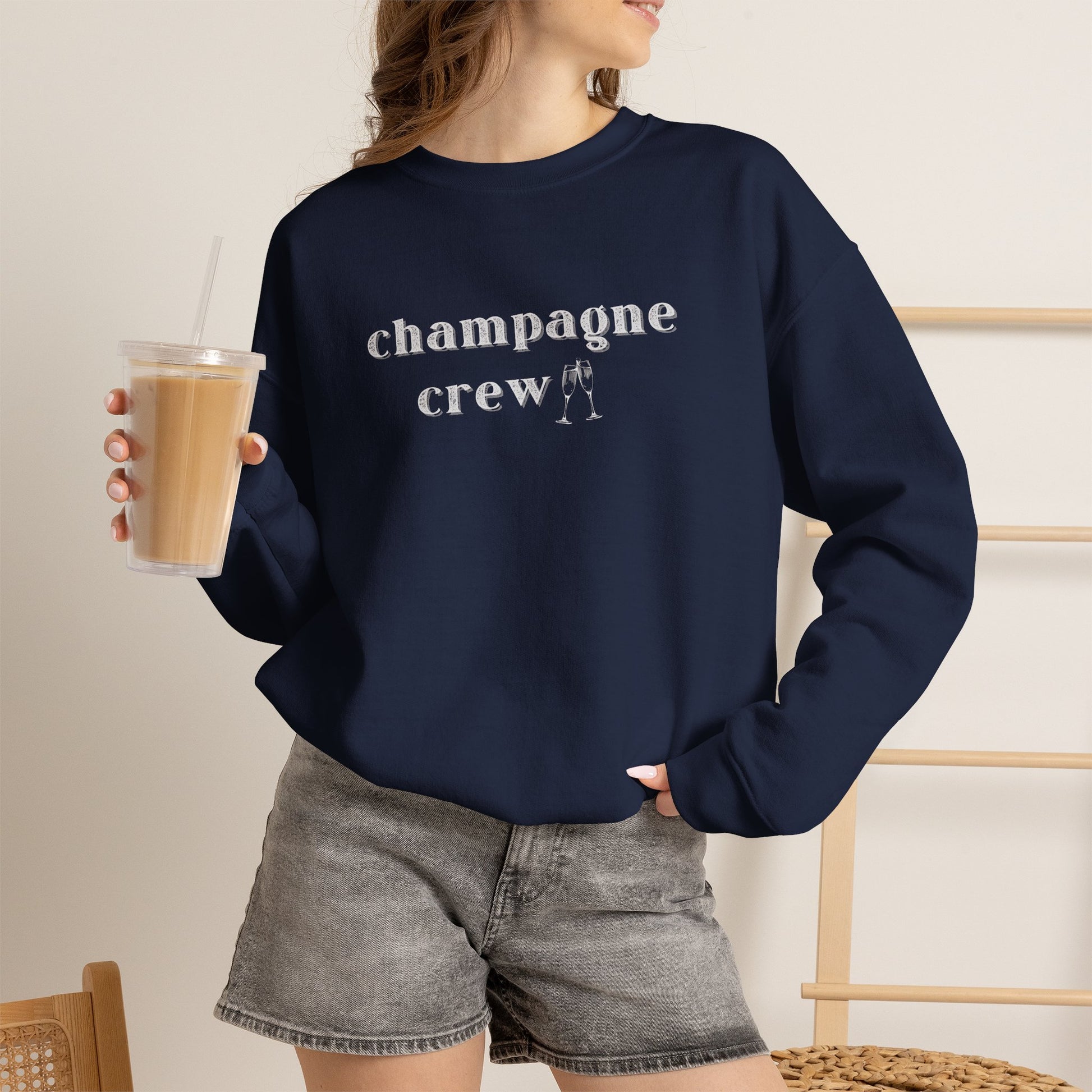 Woman wearing a navy graphic sweatshirt with text 'champagne crew' in white letters holding an iced coffee