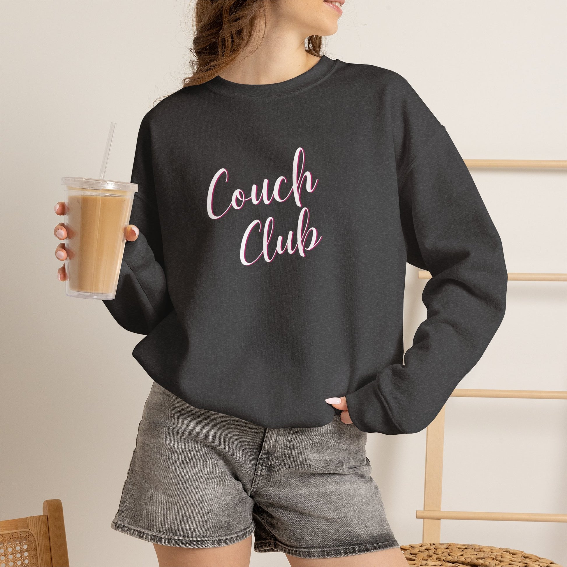 A woman wearing a dark charcoal gray crewneck sweatshirt with 'Couch Club' printed in pink script across the chest, paired with gray denim shorts and holding an iced coffee.