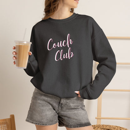 A woman wearing a dark charcoal gray crewneck sweatshirt with 'Couch Club' printed in pink script across the chest, paired with gray denim shorts and holding an iced coffee.