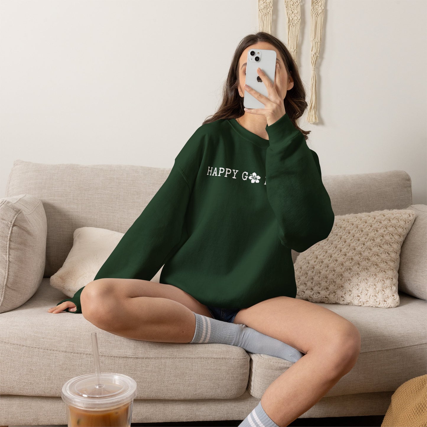 Person wearing a green sweatshirt with a graphic design ʻHAPPY GO LUCKYʻ, sitting on a couch in a living room.