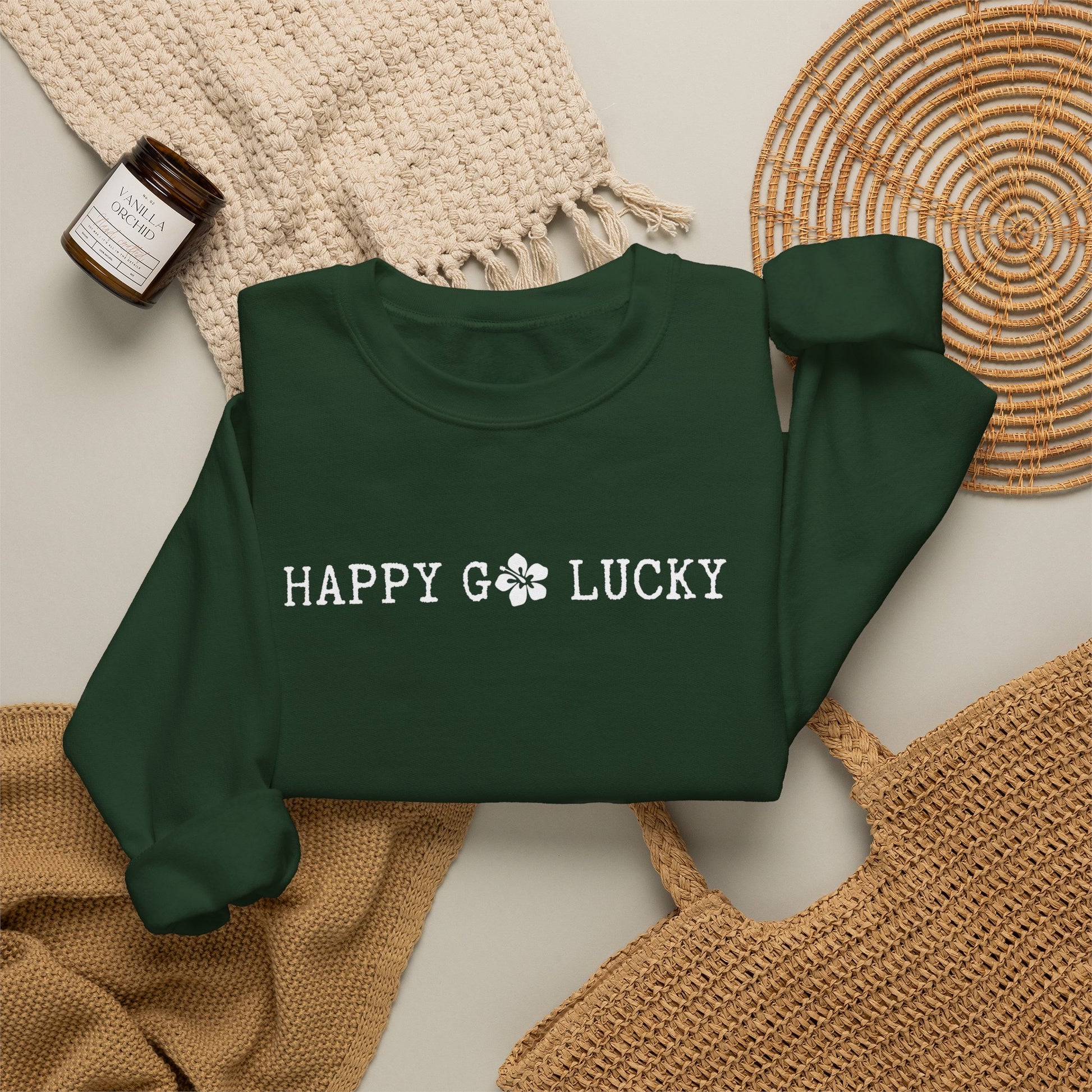 A dark green crewneck sweatshirt with 'HAPPY GO LUCKY' printed in white across the chest, styled in a flat lay with a cream knit blanket, a woven placemat, a small candle jar, and a tan woven bag on a light neutral background.