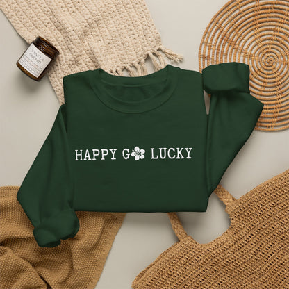 A dark green crewneck sweatshirt with 'HAPPY GO LUCKY' printed in white across the chest, styled in a flat lay with a cream knit blanket, a woven placemat, a small candle jar, and a tan woven bag on a light neutral background.