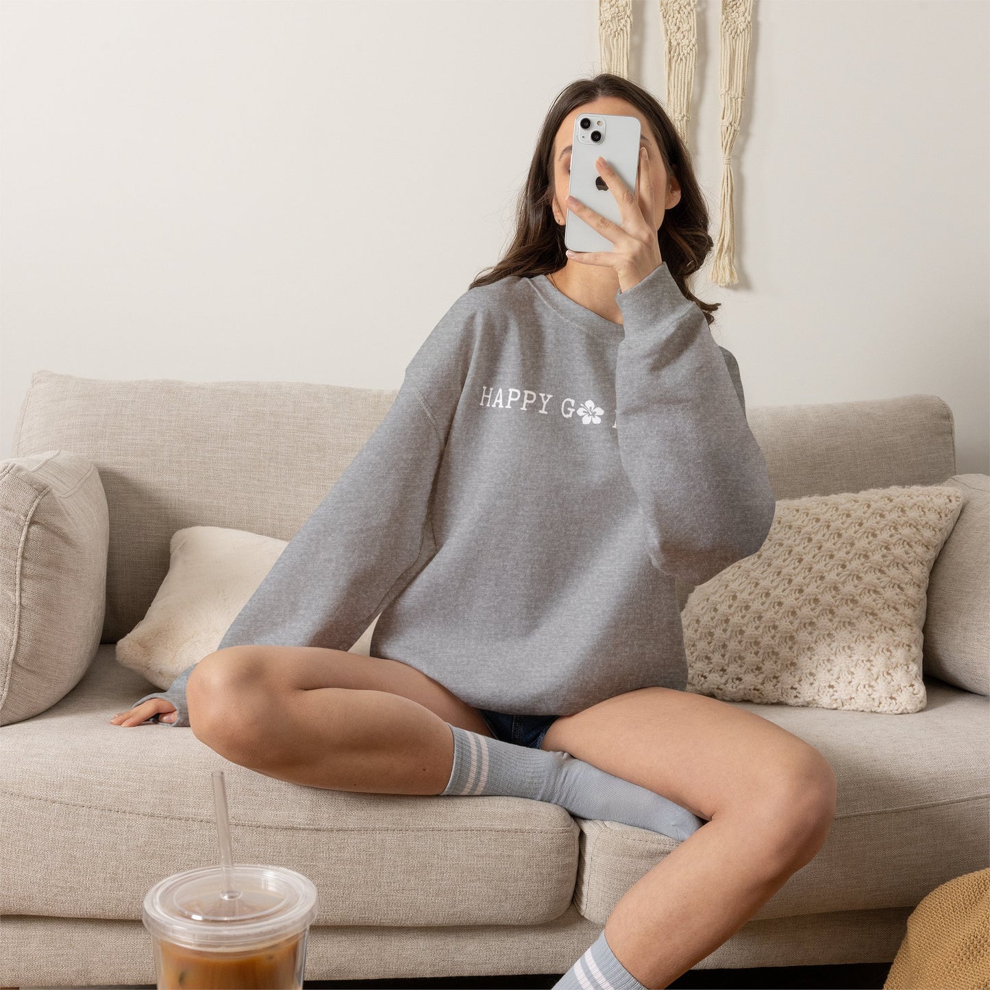 A woman takes a mirror selfie while sitting cross-legged on a beige couch, wearing a heather gray crewneck sweatshirt with 'HAPPY GO LUCKY' printed in white script across the chest.