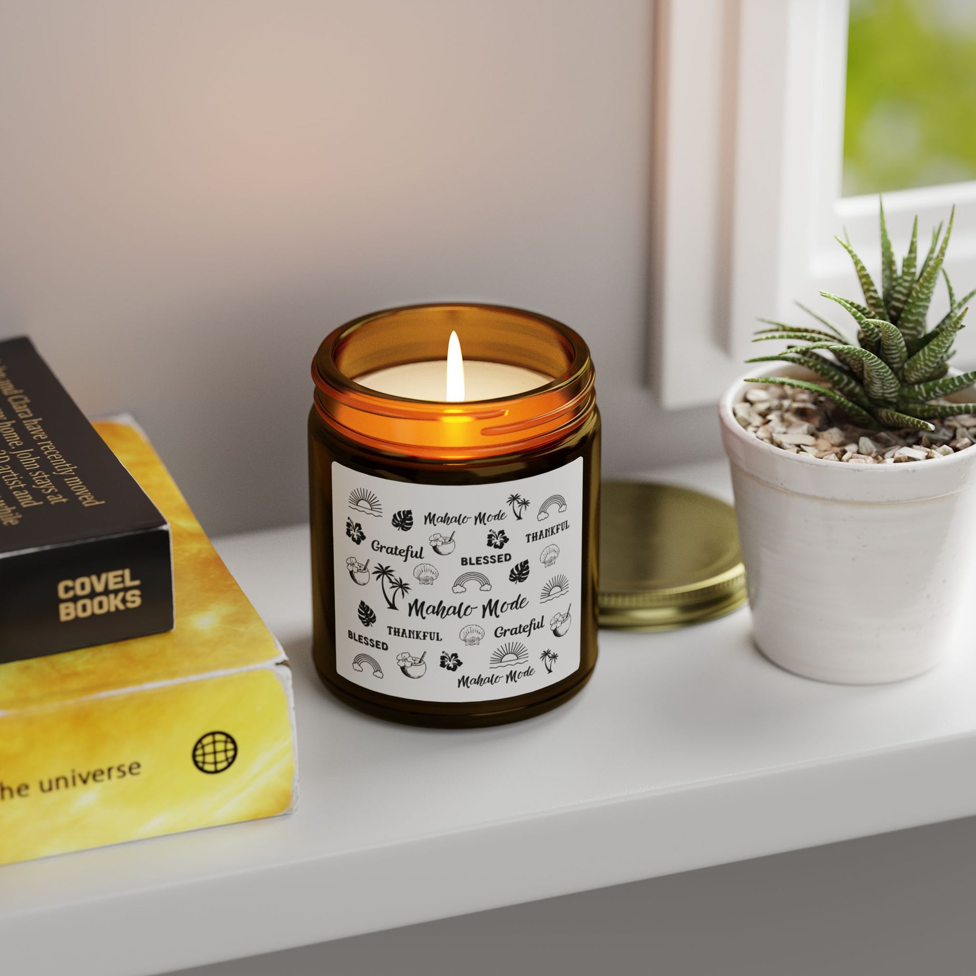 A dark amber glass jar candle with a gold metal screw-on lid and a white paper label featuring black hand-drawn style illustrations and the words 'Mahalo • Mode', 'Grateful', 'BLESSED', and 'THANKFUL' on a table with a plant and books.