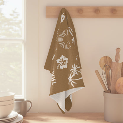 A tan kitchen towel with white tropical-themed illustrations and phrases including rainbows, palm trees, hibiscus flowers, seashells, and repeated text reading 'Grateful Mahalo Mode' and 'Blessed.' hanging from a wood peg in a kitchen