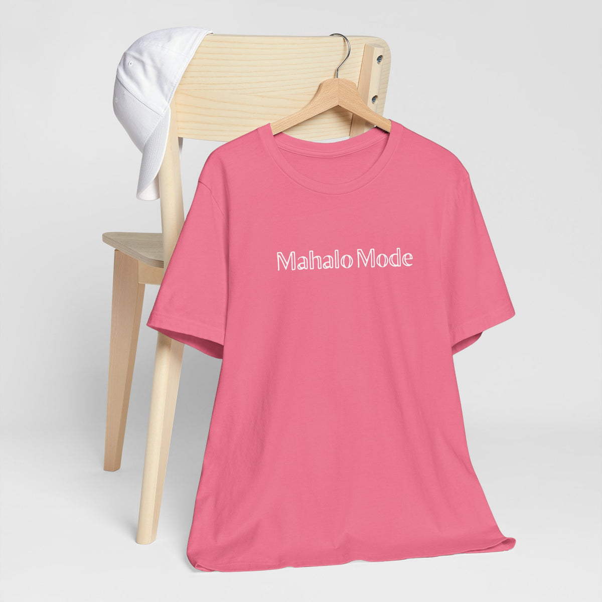 A Mahalo Mode graphic tee in charity pink with white outlined lettering, Global Village Kailua original design, hanging from a wooden chair.