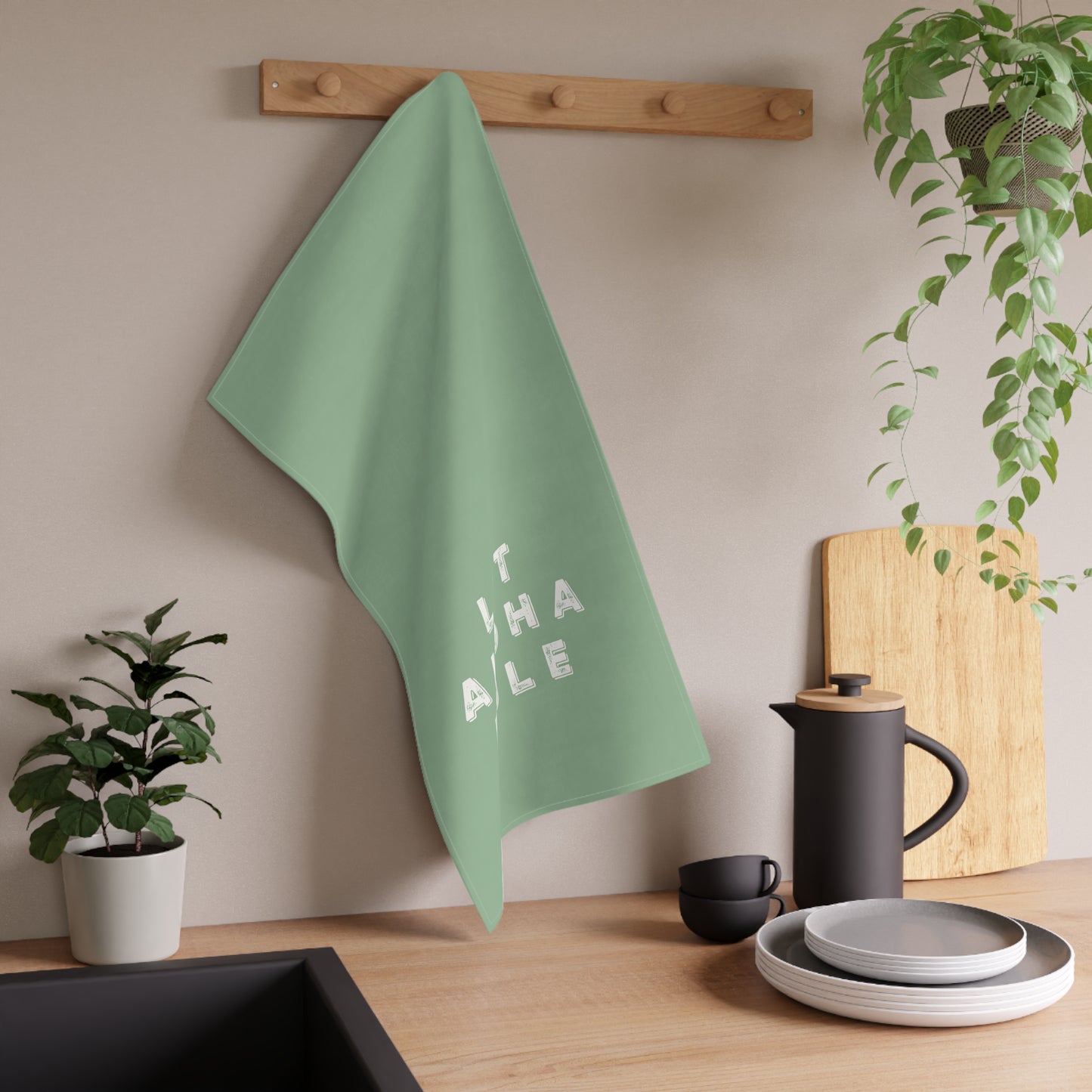 A sage green kitchen towel with the word 'LET ALOHA RULE' in white block letters hanging from a wooden peg on a kitchen wall. The scene is styled with kitchen props including a black coffee pot, plates, a cutting board, and potted plants.