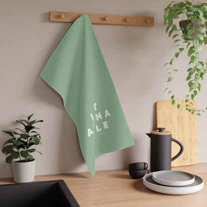 A sage green kitchen towel with the word 'LET ALOHA RULE' in white block letters hanging from a wooden peg on a kitchen wall. The scene is styled with kitchen props including a black coffee pot, plates, a cutting board, and potted plants.