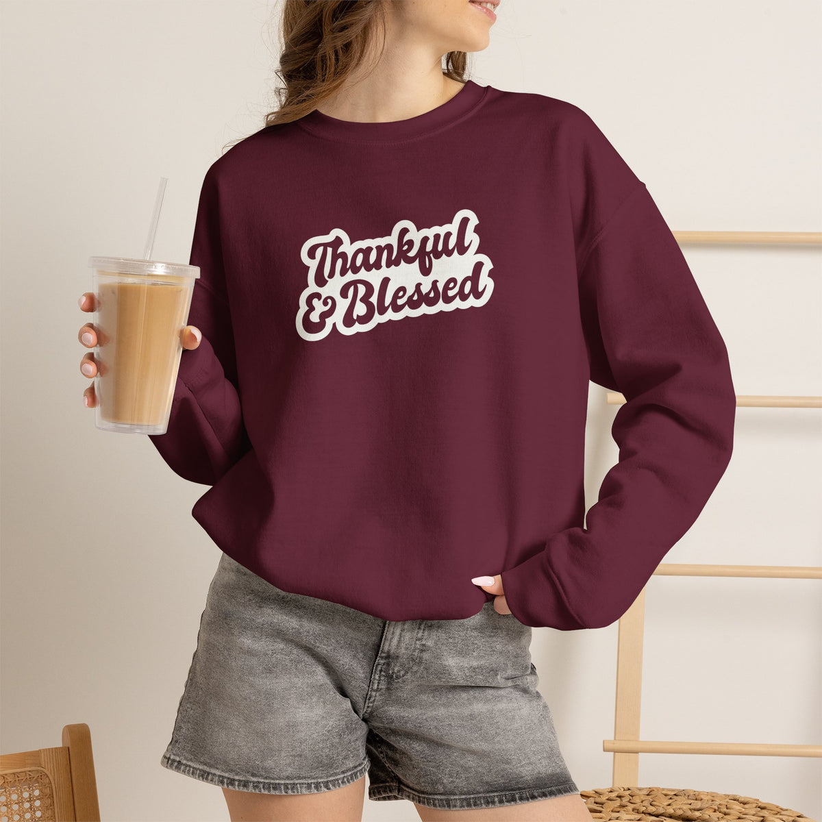 A woman wearing a maroon crewneck sweatshirt with a white graphic that reads 'Thankful & Blessed'. She is holding a iced coffee and wearing gray denim shorts in a brightly lit indoor setting.