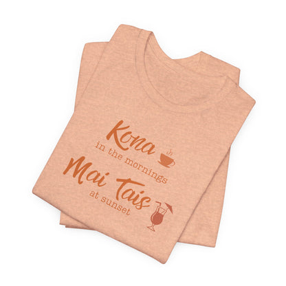 Two folded heather peach t-shirts stacked on a white background, featuring the text 'Kona in the mornings Mai Tais at sunset' in a terracotta orange font with a coffee cup and cocktail glass graphic.
