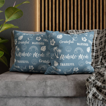 Two blue throw pillows with white tropical-themed text and illustrations, styled on a gray sofa with a plaid throw blanket and a wooden slatted wall background.