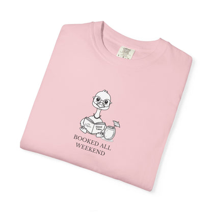 A folded light pink crewneck t-shirt featuring a cartoon graphic of a nene goose reading a book next to a cocktail glass, with the text 'BOOKED ALL WEEKEND' printed below.