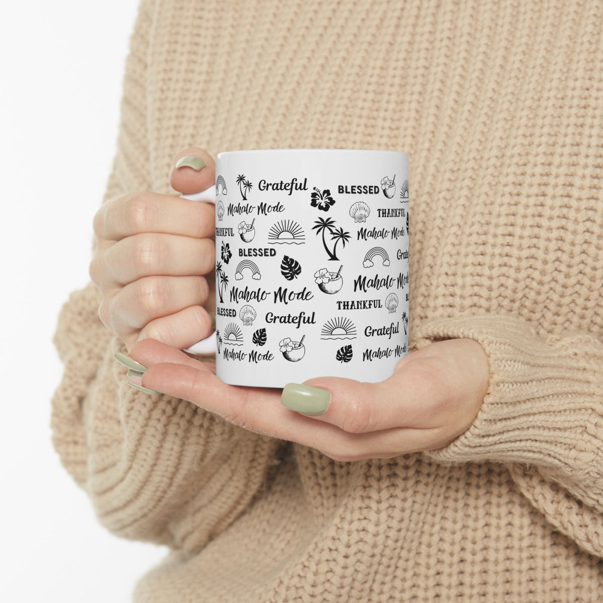 A person in a beige knit sweater holds a white ceramic mug with black tropical-themed text and illustrations, including 'Grateful', 'Blessed', 'Thankful', and 'Mahalo Mode'.