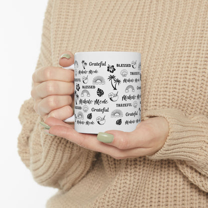 A person in a beige knit sweater holds a white ceramic mug with black tropical-themed text and illustrations, including 'Grateful', 'Blessed', 'Thankful', and 'Mahalo Mode'.