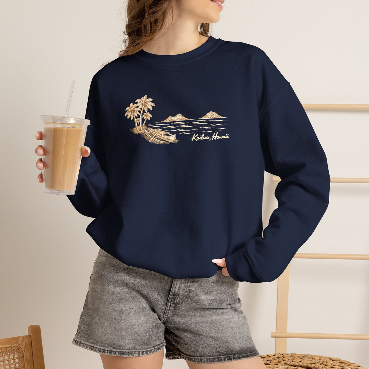 A woman wearing a navy blue crewneck sweatshirt with a beige graphic of palm trees, mountains, and the text 'Kailua Hawaii' on the front. She is holding a iced coffee and wearing gray denim shorts in a brightly lit indoor setting.