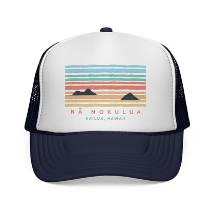 A white and navy blue trucker hat featuring a colorful graphic of a sunset over mountains with the text 'NÁ MOKULUA' and 'KAILUA, HAWAII'.