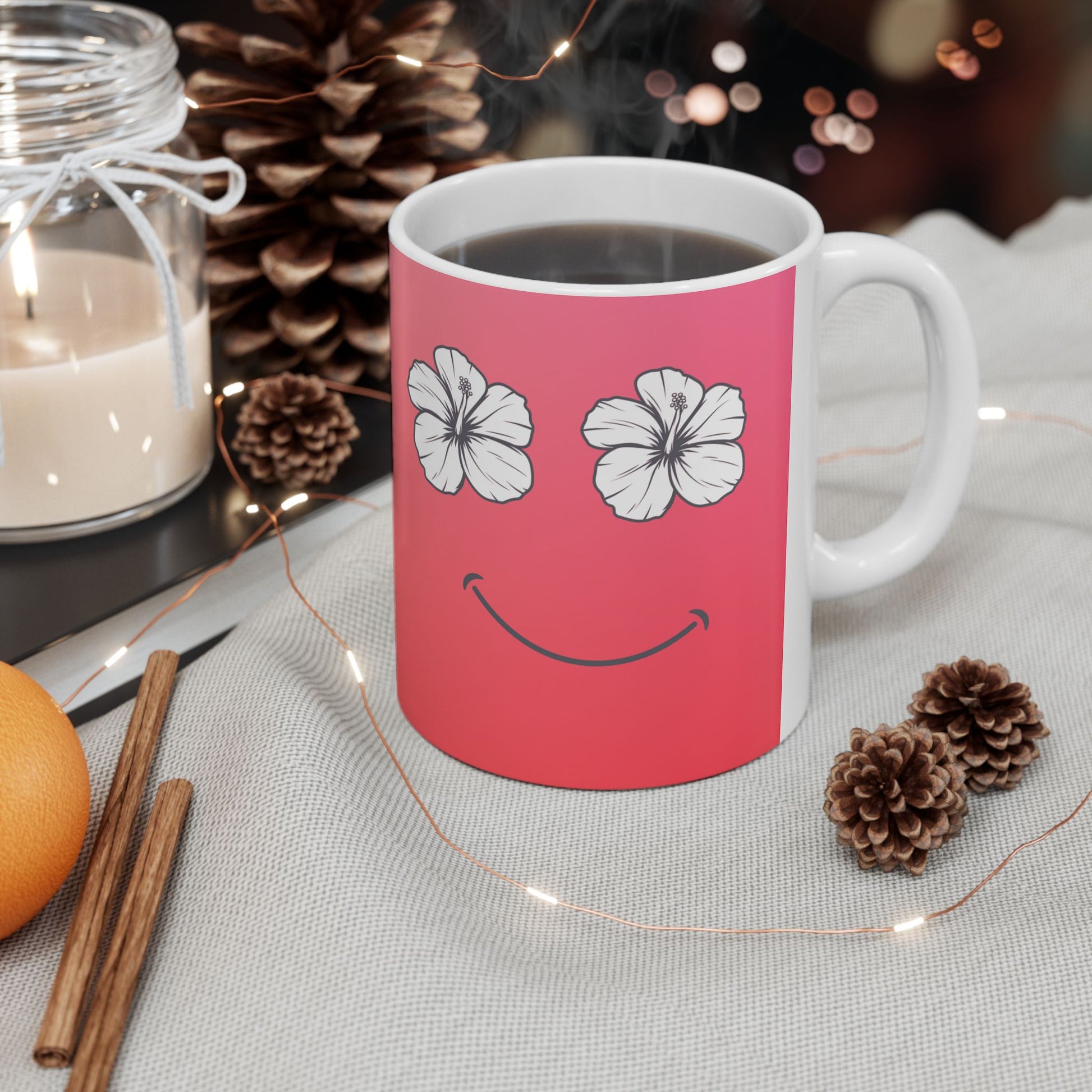 A white ceramic mug with a pink front panel featuring a smiley face made of white hibiscus flowers and a curved smile, filled with a dark beverage. The mug sits on a light fabric surface surrounded by pinecones, a candle jar, cinnamon sticks, and fairy lights.