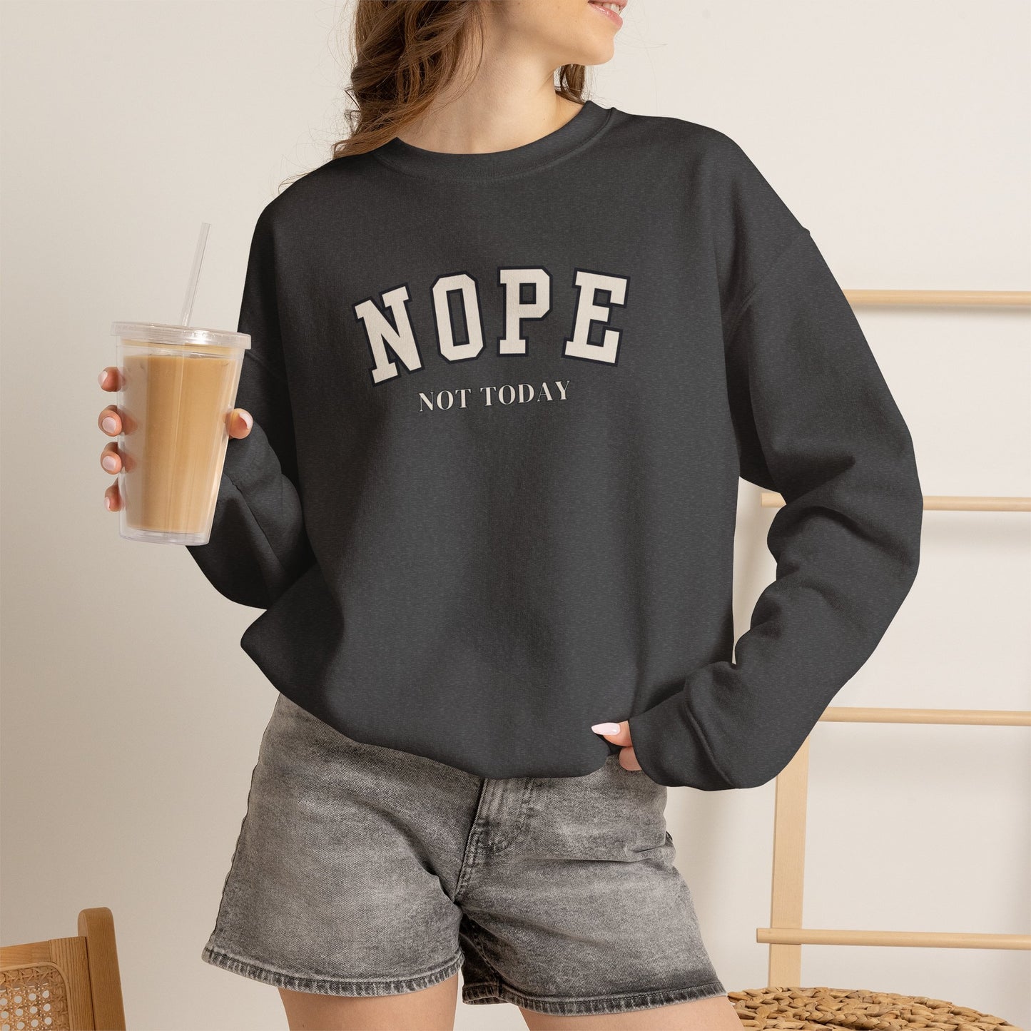 A woman wearing a dark charcoal gray crewneck sweatshirt with the text 'NOPE NOT TODAY' printed in white on the front, paired with gray denim shorts and holding an iced coffee.