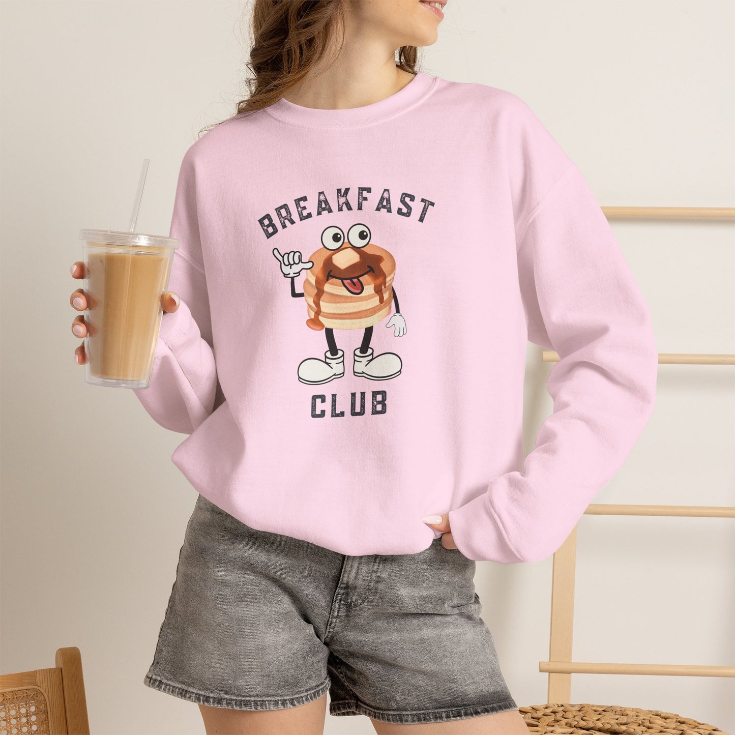 A woman wearing a light pink crewneck sweatshirt with a cartoon breakfast graphic that reads 'BREAKFAST CLUB'. She is holding an iced coffee and wearing gray denim shorts in a brightly lit indoor setting.