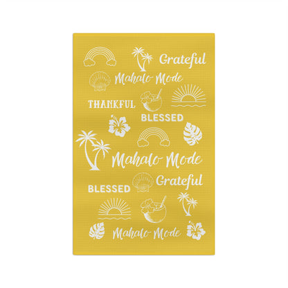 A yellow waffle-weave kitchen towel with white tropical-themed text 'mahalo mode' 'thankful' 'blessed' 'grateful' and illustrations, hanging from a clothesline with wooden clips against a white background.