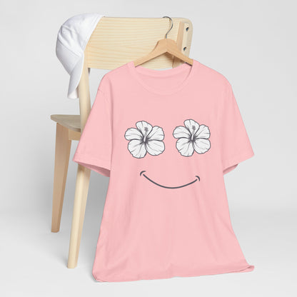 A light pink short-sleeve t-shirt with a graphic of two white hibiscus flowers forming eyes and a black smiley mouth, hanging on a wooden hanger from a light wood chair.