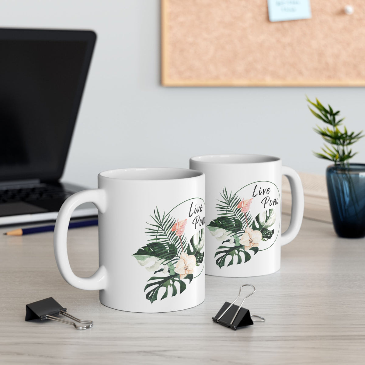 Two white ceramic mugs with a tropical leaf and peach flower graphic and the text 'Live Pono' printed on them, displayed on a light wood desk with office props in the background.