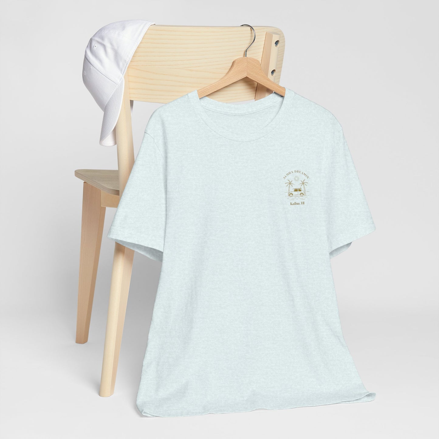 A light heather blue crewneck t-shirt with a small gold 'Aloha Dreamin Kailua Hawaii' design on the left chest, displayed on a wooden hanger draped over a light wood chair with a white shirt hanging off the backrest, against a neutral light gray background.
