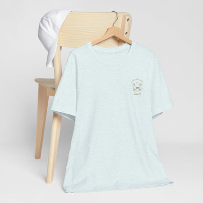 A light heather blue crewneck t-shirt with a small gold 'Aloha Dreamin Kailua Hawaii' design on the left chest, displayed on a wooden hanger draped over a light wood chair with a white shirt hanging off the backrest, against a neutral light gray background.