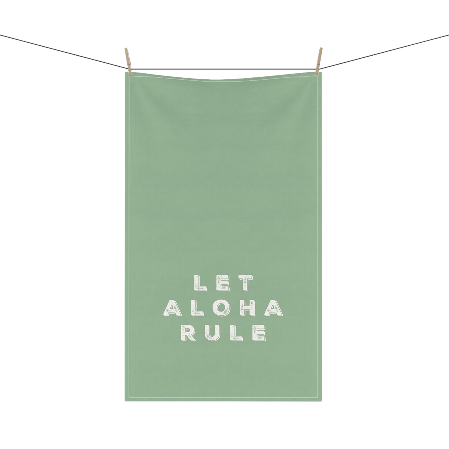 A sage green kitchen towel with the phrase 'LET ALOHA RULE' printed in white textured block letters, hanging from a clothesline with two wooden clips against a solid white background.