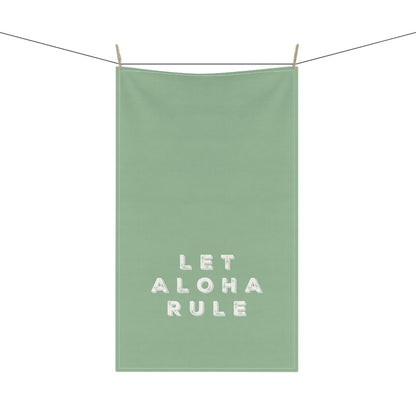 A sage green kitchen towel with the phrase 'LET ALOHA RULE' printed in white textured block letters, hanging from a clothesline with two wooden clips against a solid white background.