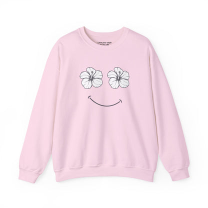 A light pink crewneck sweatshirt featuring a graphic of two white hibiscus flowers forming eyes and a simple black smiley curve below, displayed on a clean white background.