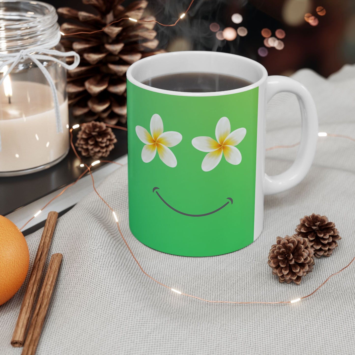 A white ceramic mug with a green front panel featuring two white plumeria flowers and a smiley face, filled with a hot beverage and surrounded by cozy holiday props like pinecones, a candle, cinnamon sticks, and fairy lights.