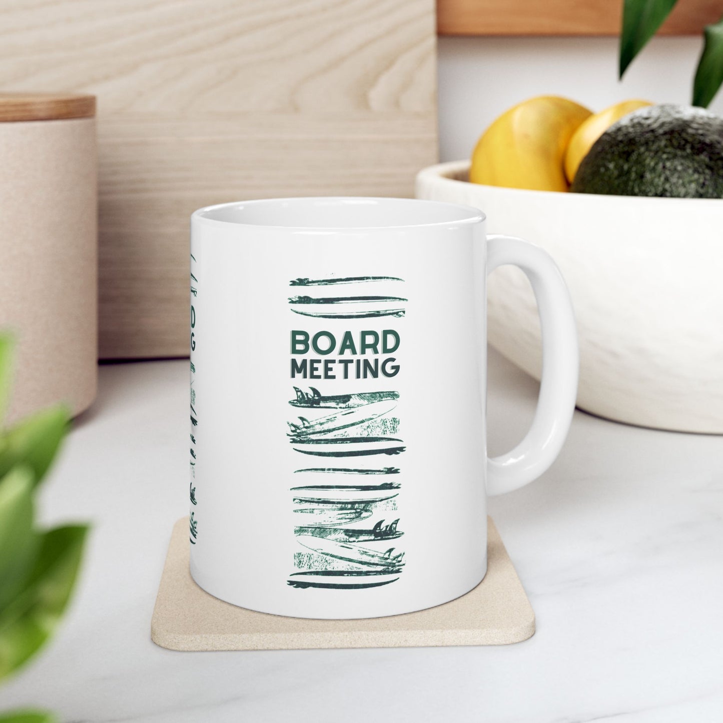 A white ceramic mug with a dark green 'BOARD MEETING' text design and horizontal brushstroke graphics, sitting on a light wood coaster on a white marble countertop with a blurred kitchen background.
