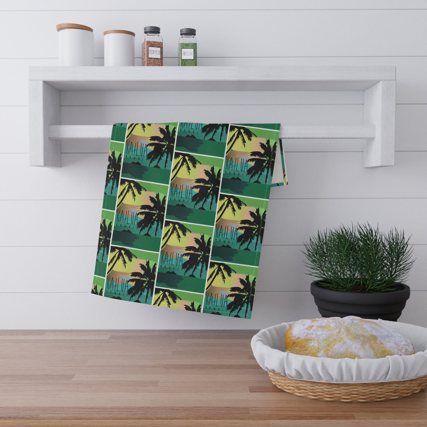 A kitchen towel with a tropical palm tree sunset pattern 'Kailua Hawaii' hangs from a white wall-mounted rack in a bright kitchen setting.